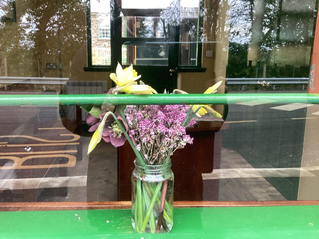 Through the Waiting Room window Welcoming flowers as often provided in the past at stations, the 1930s Art Deco furniture, the 1025 train to Exeter Central with about 40 passengers, a deserted Downside platform bench and in the distance the former Goods Shed, now a Youth Hostel.brPhotographer A visitor to OkehamptonbrDate taken 06032023